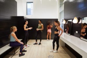 photo of five women in workout clothes talking in gym locker room with black tile walls.
