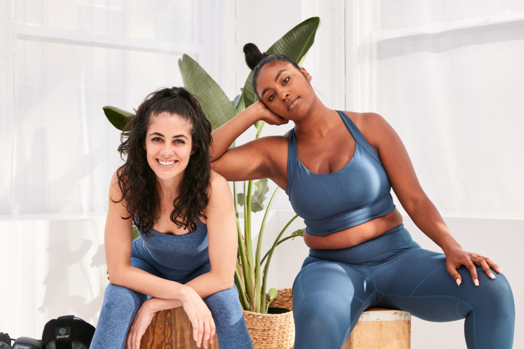 Photo of two women in blue workout gear sitting in front of white windows 